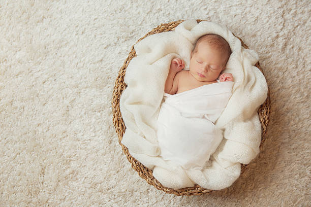 Newborn in White Swaddle Blanket sleeping in Basket. One month Baby lying on back on White Carpet Background.