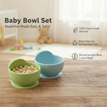Two baby bowls, one green and one blue, on a wooden surface with a baby's crib and toys in the background.