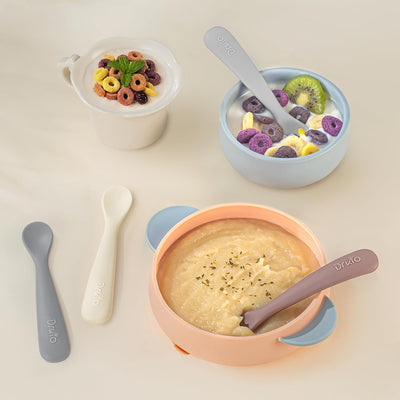 Assorted baby feeding bowls and spoons with different food items on a light background