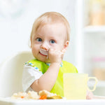 Baby sitting in a high chair with a bib, surrounded by food and a cup.