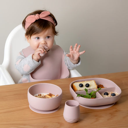 Baby sitting at a table with pink tableware, eating a meal.