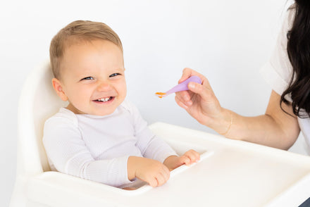 Child sitting in a high chair with a spoon held by an adult, on a white background