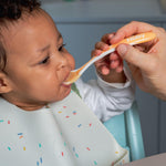 Child being fed with a spoon by an adult, wearing a bib with colorful sprinkles.