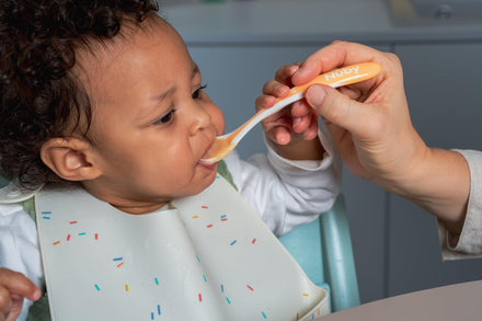 Child being fed with a spoon by an adult, wearing a bib with colorful sprinkles.