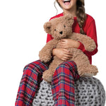 Child in red shirt and plaid pants holding a teddy bear on a gray bean bag chair.