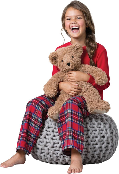 Child in red shirt and plaid pants holding a teddy bear on a gray bean bag chair.